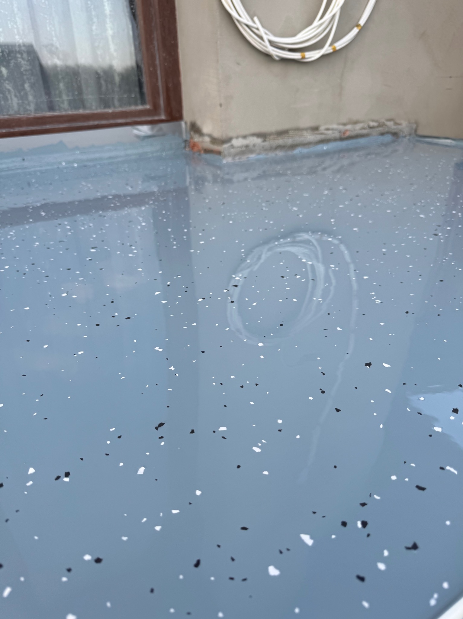 Błękitna, błyszcząca powierzchnia z czarno-białymi drobinkami, świeżo wylana, odbija światło okna i zwisający kabel. Gładka, jednolita struktura.