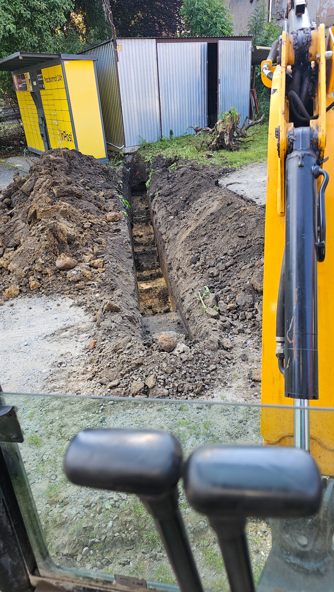 Wąski wykop w ziemi, widziany z kabiny koparki na tle Paczkomatu i blaszanej budki. Świeżo wykopany rów z równymi ścianami.