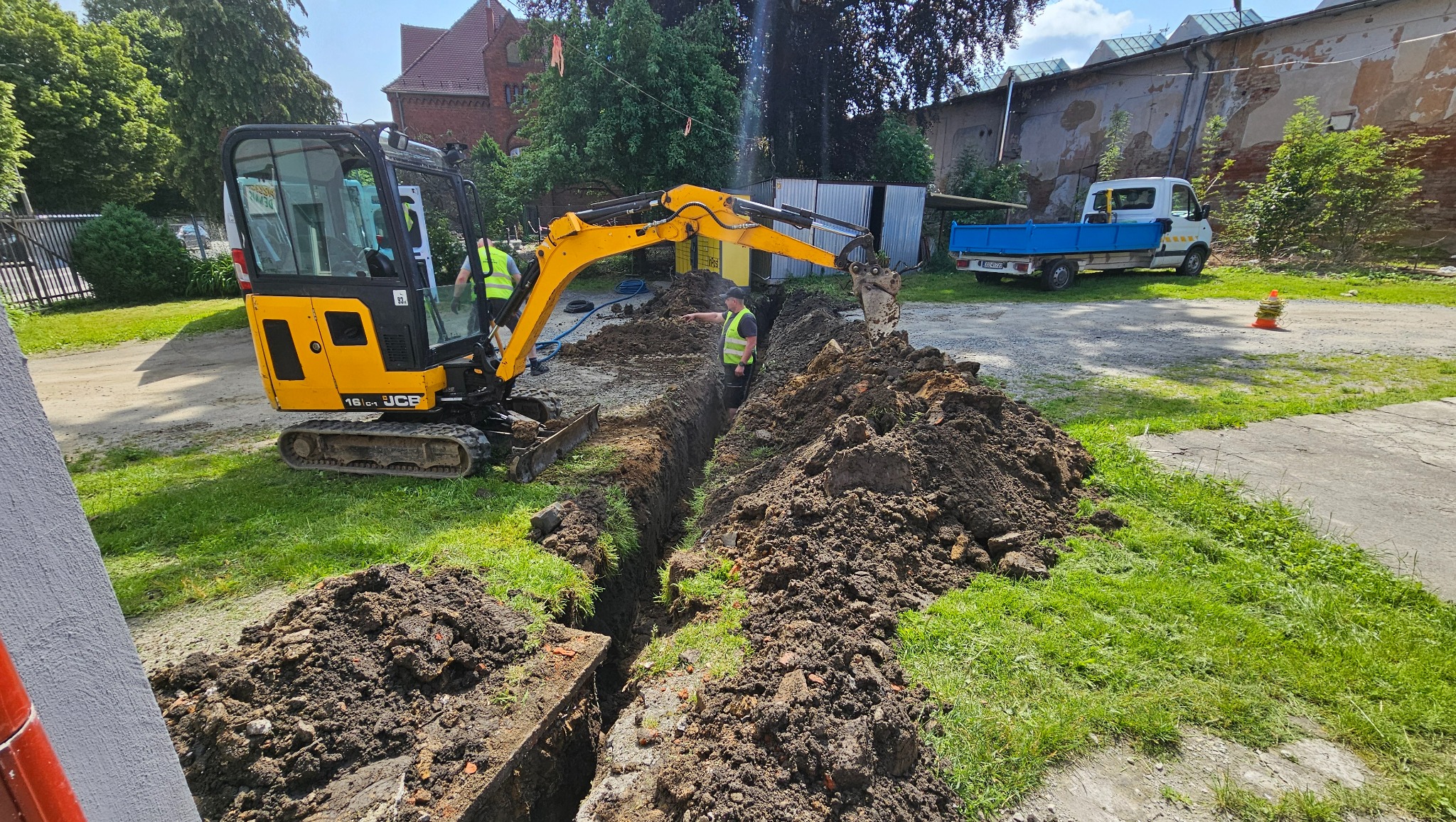 Żółta mini koparka JCB podczas wykopu wąskiego rowu w Pieszycach. Dwóch pracowników w kamizelkach odblaskowych nadzoruje prace. W tle budynki i biała ciężarówka.
