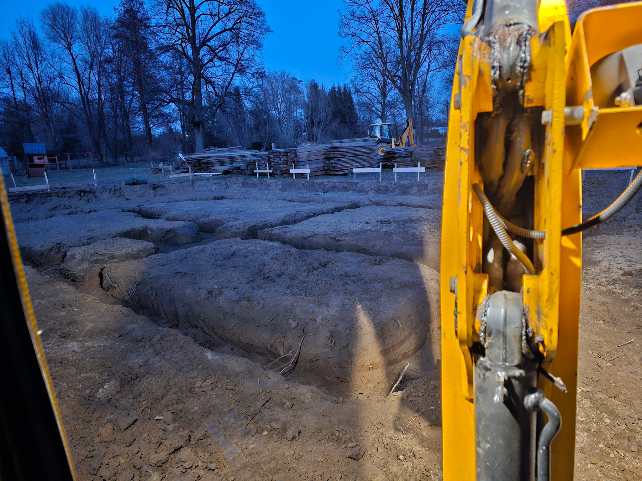 Wykopy pod fundamenty, widok z perspektywy operatora koparki. W tle zapas drewna budowlanego i oddalona koparka. Wyraźne ślady pracy maszyny.