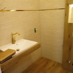 Mariusz haberman - Wykończona łazienka z umywalką na drewnianym blacie, złota armatura i dekoracyjne listwy. Płytki ścienne z delikatnym wzorem fal. Drewniana podłoga.