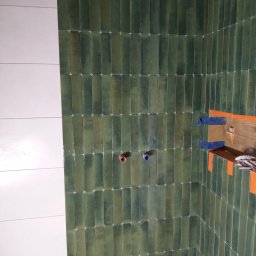 Mariusz haberman - Ukończona ściana łazienki z zielonymi, podłużnymi płytkami i białymi fugami. Widoczne przygotowane punkty instalacji wodnej i fragment zabezpieczonej półki.