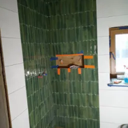 Ściana z zielonych, wąskich płytek w łazience, z wnęką zabezpieczoną taśmą. Część ściany wykończona białymi płytkami. Na podłodze drewnopodobne panele.