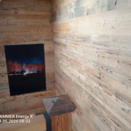 Mariusz haberman - Drewniana boazeria na ścianach w rogu pomieszczenia, z obrazem na ścianie po lewej stronie. Widoczny fragment schodów i drewniany słupek.