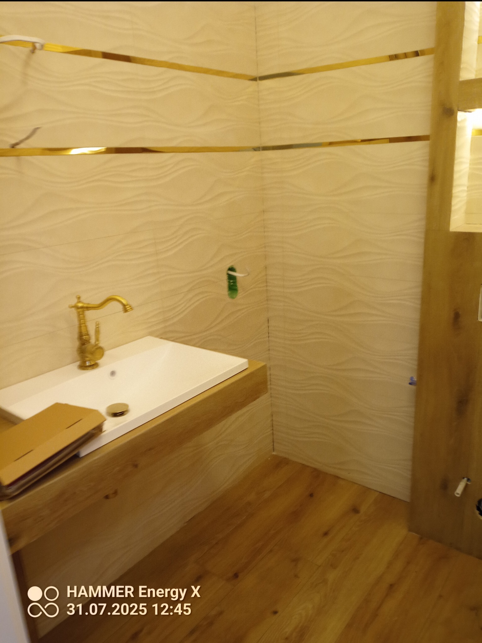 Wykończona łazienka z umywalką na drewnianym blacie, złota armatura i dekoracyjne listwy. Płytki ścienne z delikatnym wzorem fal. Drewniana podłoga.