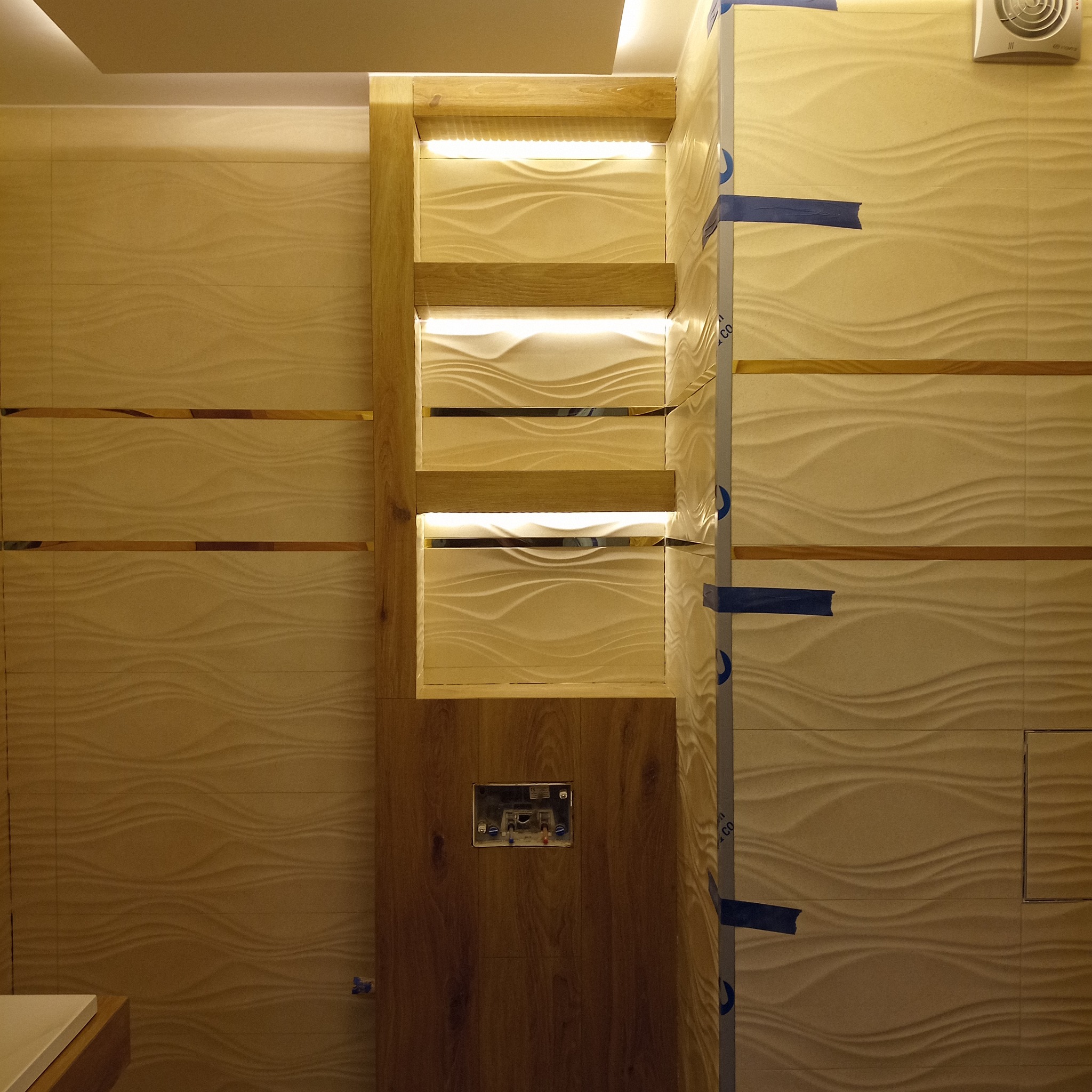 Wykończona łazienka z podświetlanymi półkami z drewna i dekoracyjnymi płytkami w odcieniach beżu. Widoczny fragment umywalki.