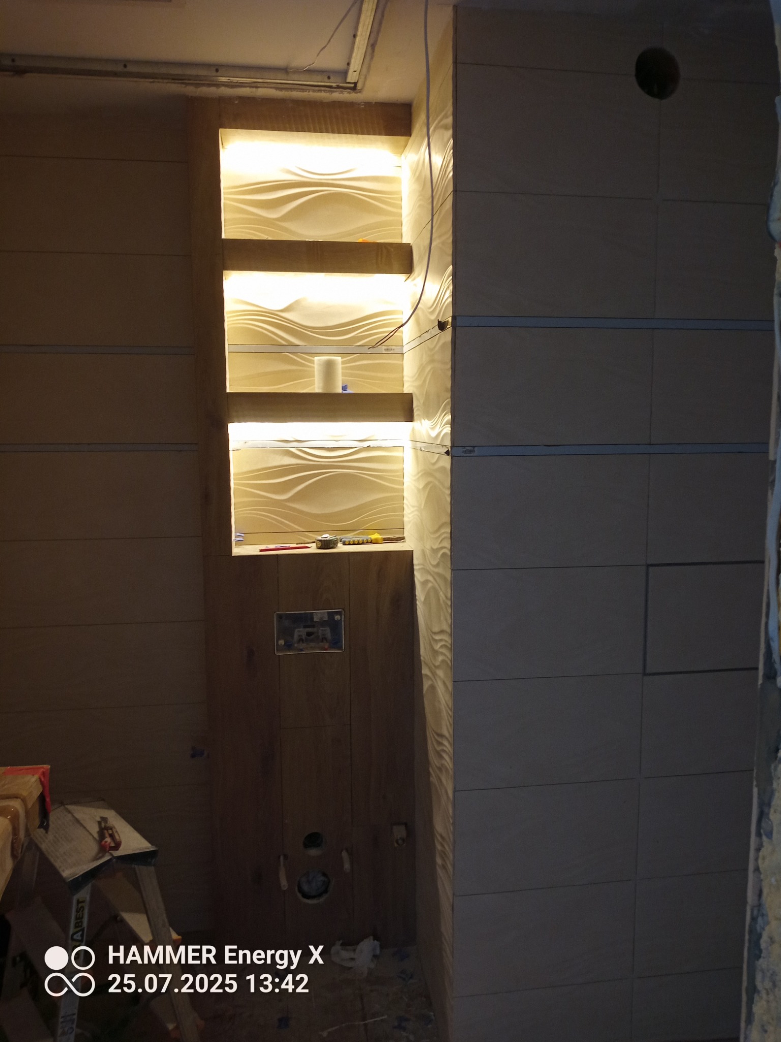 Płytki imitujące drewno na ścianie z wnęką oświetloną LED, w której znajdują się dekoracyjne panele 3D. Widoczne narzędzia i drabina, sugerujące trwający remont.