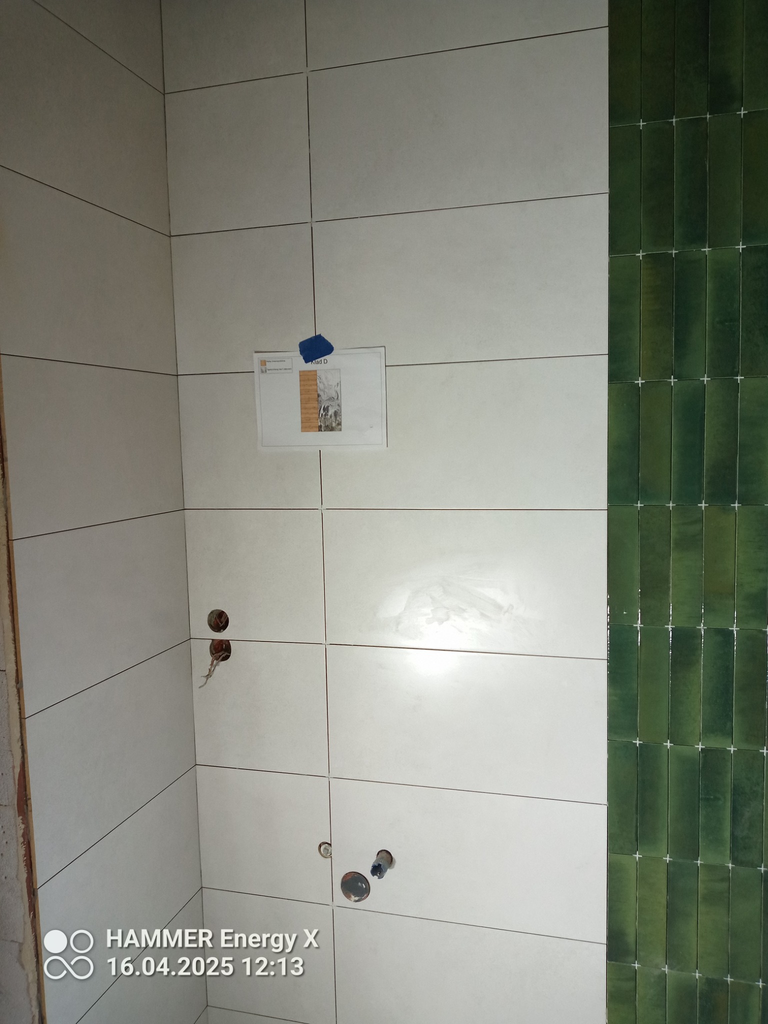 Ściana w trakcie wykańczania w Giżycku: białe płytki ceramiczne zestawione z pionowym pasem zielonych, wąskich płytek. Widoczne otwory na armaturę i dokumentacja.