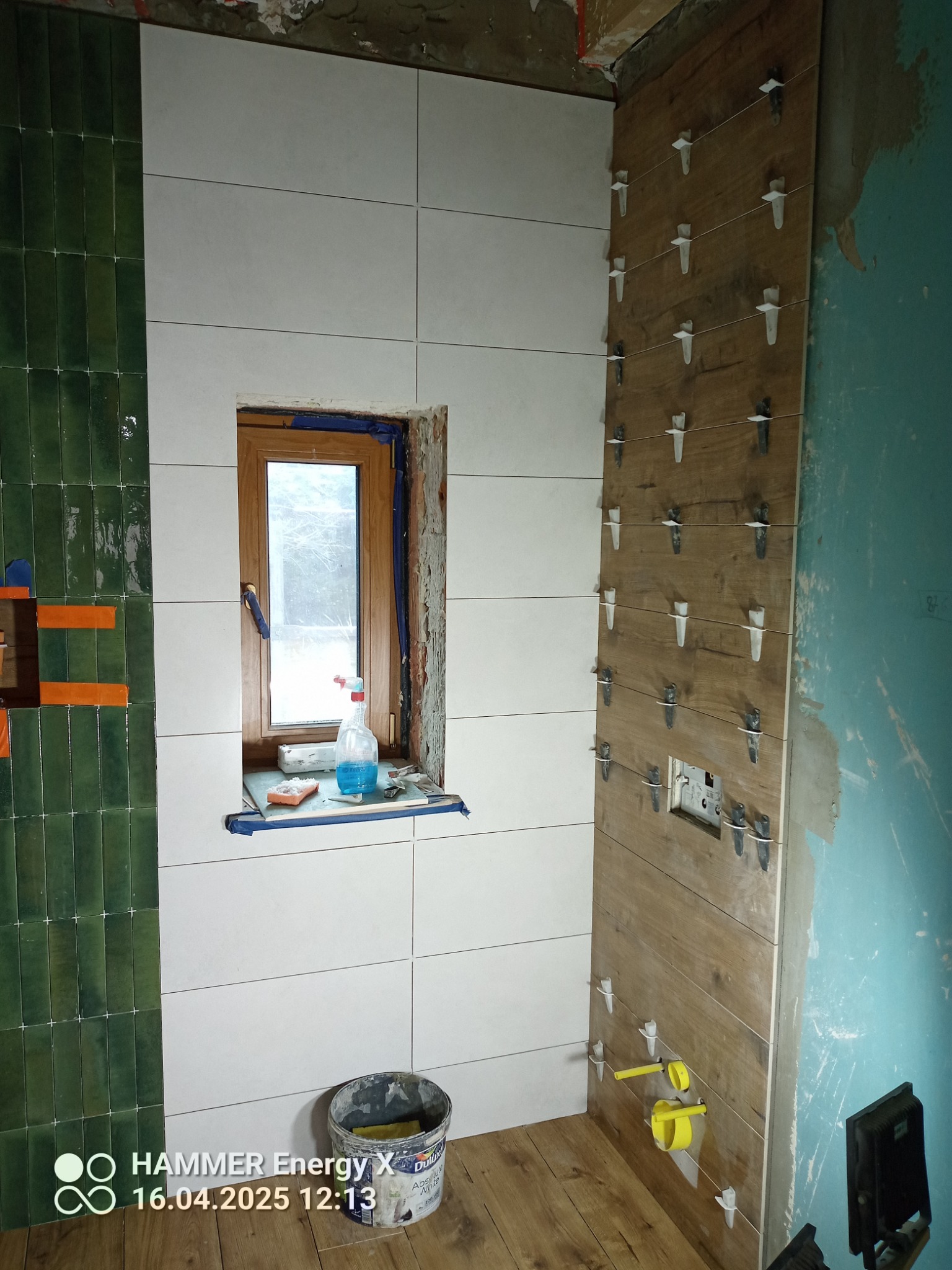 Układanie płytek w łazience: zielone, białe i drewnopodobne płytki na ścianach, widoczne krzyżyki dystansowe, okno z akcesoriami i wiadro z klejem na podłodze.