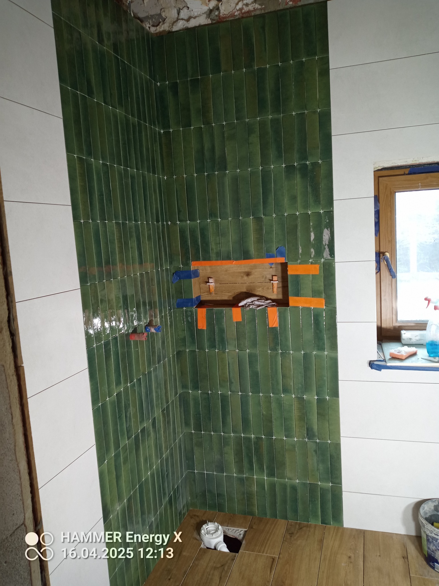 Ściana z zielonych, wąskich płytek w łazience, z wnęką zabezpieczoną taśmą. Część ściany wykończona białymi płytkami. Na podłodze drewnopodobne panele.