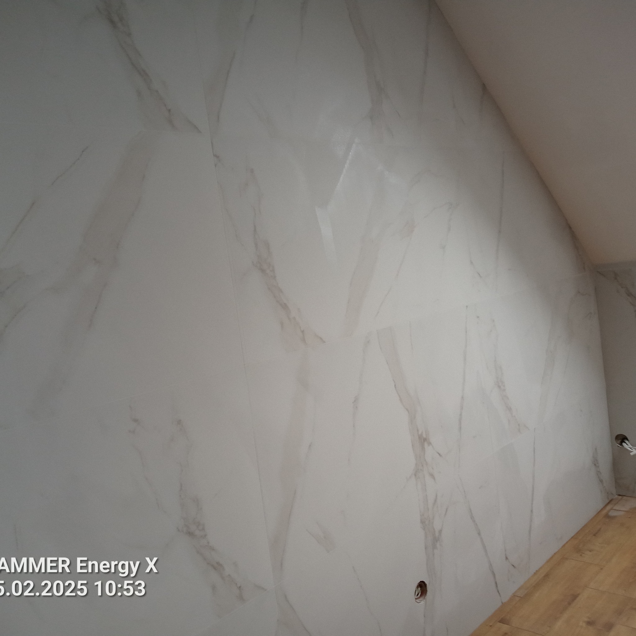 Ściana w łazience wyłożona białymi płytkami imitującymi marmur, z widocznymi otworami na instalacje. Podłoga wyłożona panelami imitującymi drewno. Ukośny sufit.