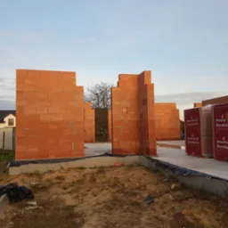 Wznoszenie ścian parteru z czerwonej ceramiki Porotherm na fundamencie. Częściowo wymurowane ściany budynku, w tle dom i błękitne niebo.