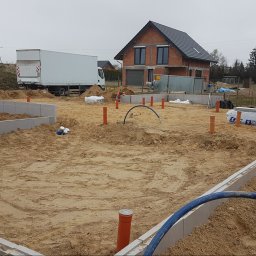 ALS Design Mateusz Pawelczyk - Wykopane i przygotowane fundamenty pod budowę domu, z widoczną konstrukcją ścian fundamentowych i wystającymi rurami kanalizacyjnymi. W tle dom w stanie surowym otwartym.