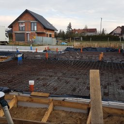 ALS Design Mateusz Pawelczyk - Zbrojenie fundamentów pod budowę domu jednorodzinnego. Widoczne szalunki, siatka zbrojeniowa i wystające rury instalacyjne. W tle dom w stanie surowym otwartym i samochód dostawczy.