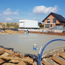 ALS Design Mateusz Pawelczyk - Wylany fundament pod dom jednorodzinny z widocznym zbrojeniem i instalacjami. W tle budynek w trakcie budowy i biała ciężarówka dostawcza.