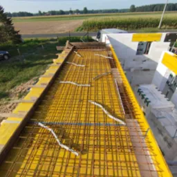 Zbrojenie stropu na żółtym szalunku, widok z góry na budowę domu jednorodzinnego z bloczków, w tle krajobraz wiejski w słoneczny dzień.