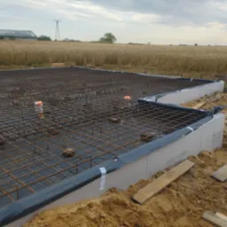 Zbrojenie fundamentów pod budowę domu jednorodzinnego, widok na siatkę zbrojeniową i szalunki, w tle pole i konstrukcja stalowa.