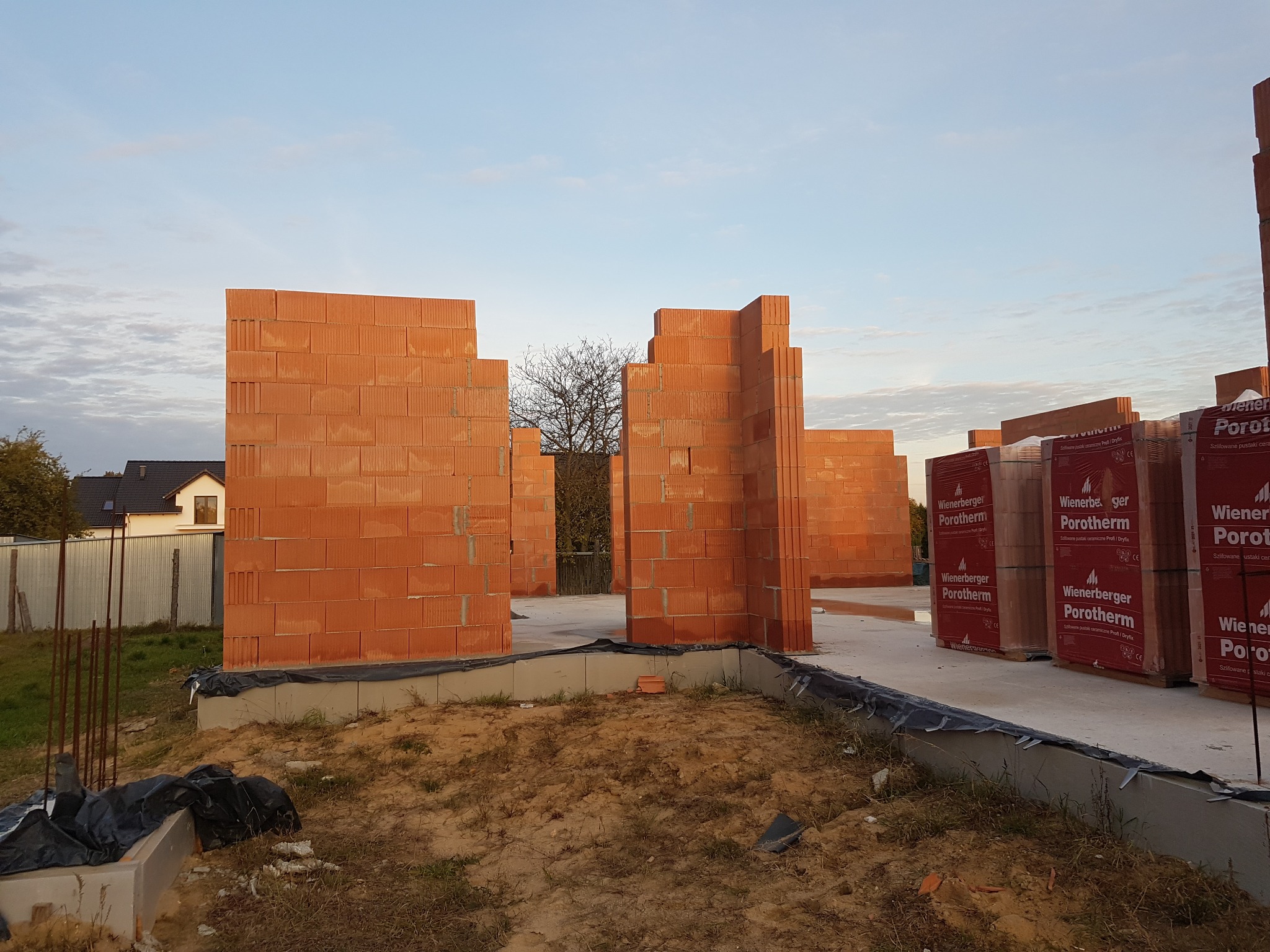 Wznoszenie ścian parteru z czerwonej ceramiki Porotherm na fundamencie. Częściowo wymurowane ściany budynku, w tle dom i błękitne niebo.