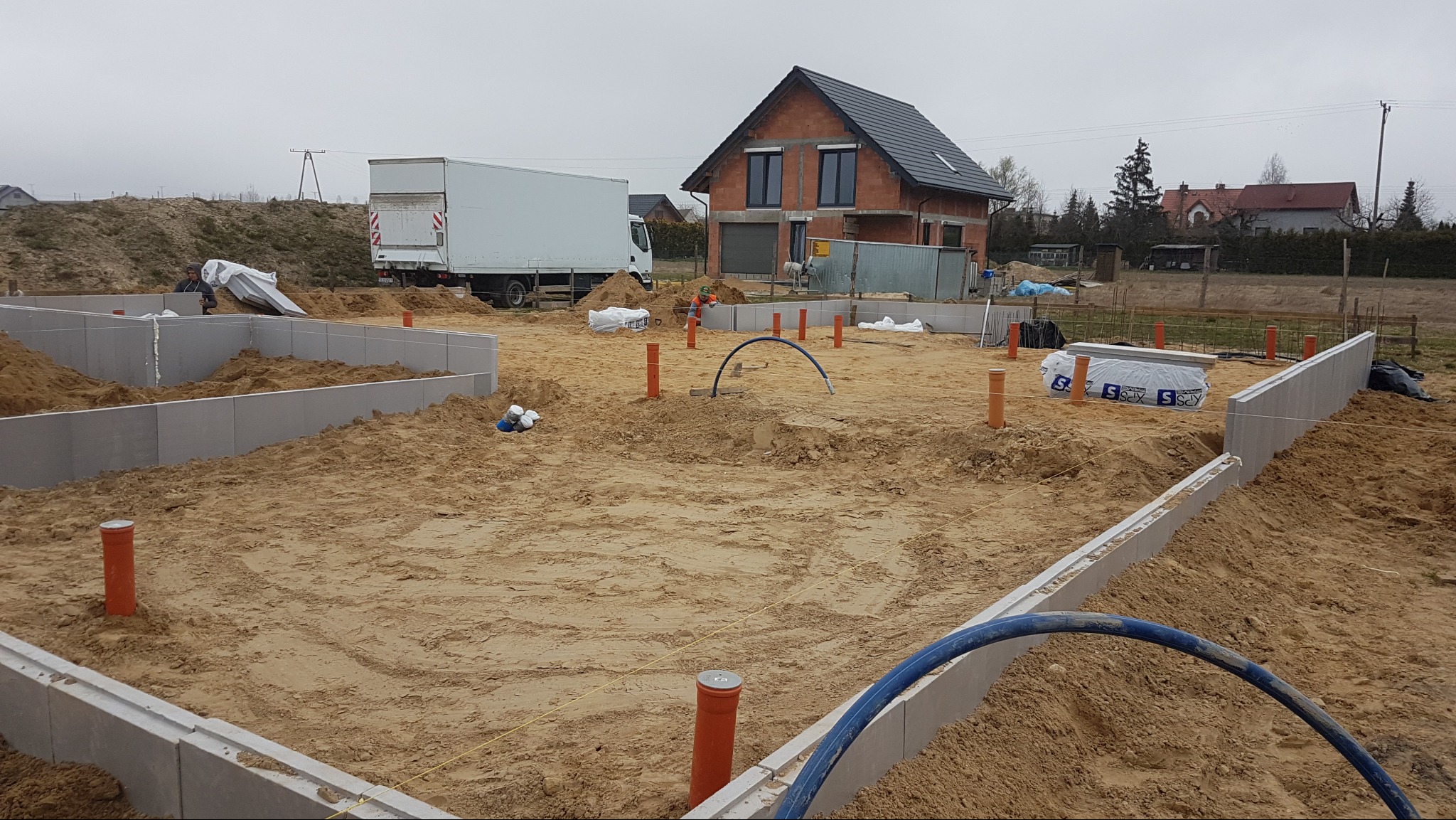 Wykopane i przygotowane fundamenty pod budowę domu, z widoczną konstrukcją ścian fundamentowych i wystającymi rurami kanalizacyjnymi. W tle dom w stanie surowym otwartym.