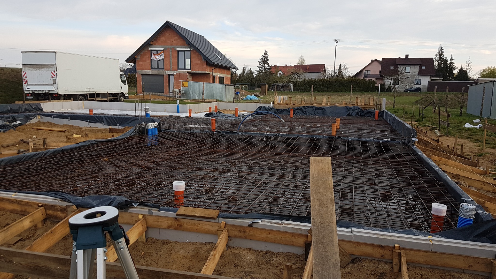Zbrojenie fundamentów pod budowę domu jednorodzinnego. Widoczne szalunki, siatka zbrojeniowa i wystające rury instalacyjne. W tle dom w stanie surowym otwartym i samochód dostawczy.