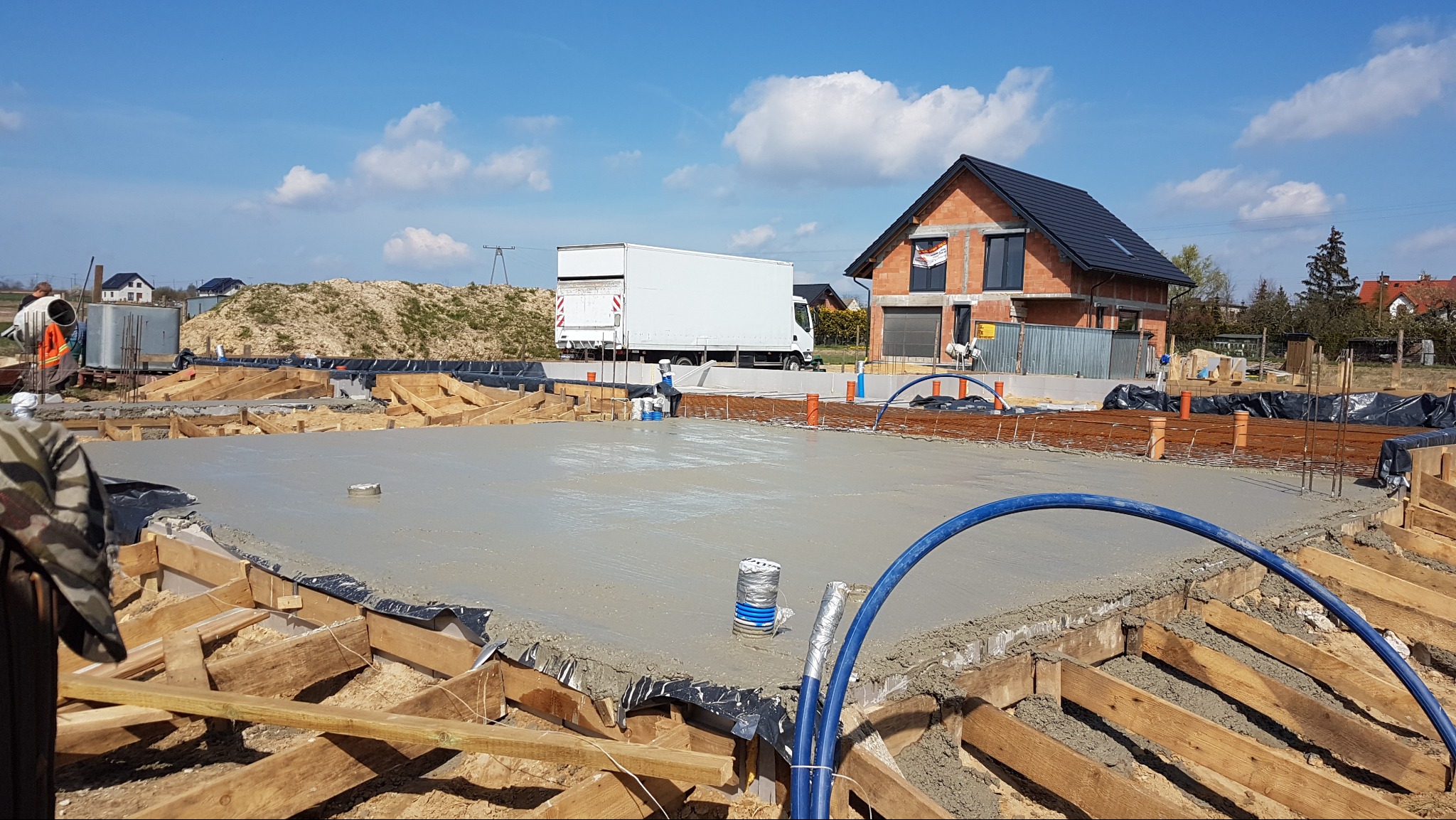 Wylany fundament pod dom jednorodzinny z widocznym zbrojeniem i instalacjami. W tle budynek w trakcie budowy i biała ciężarówka dostawcza.