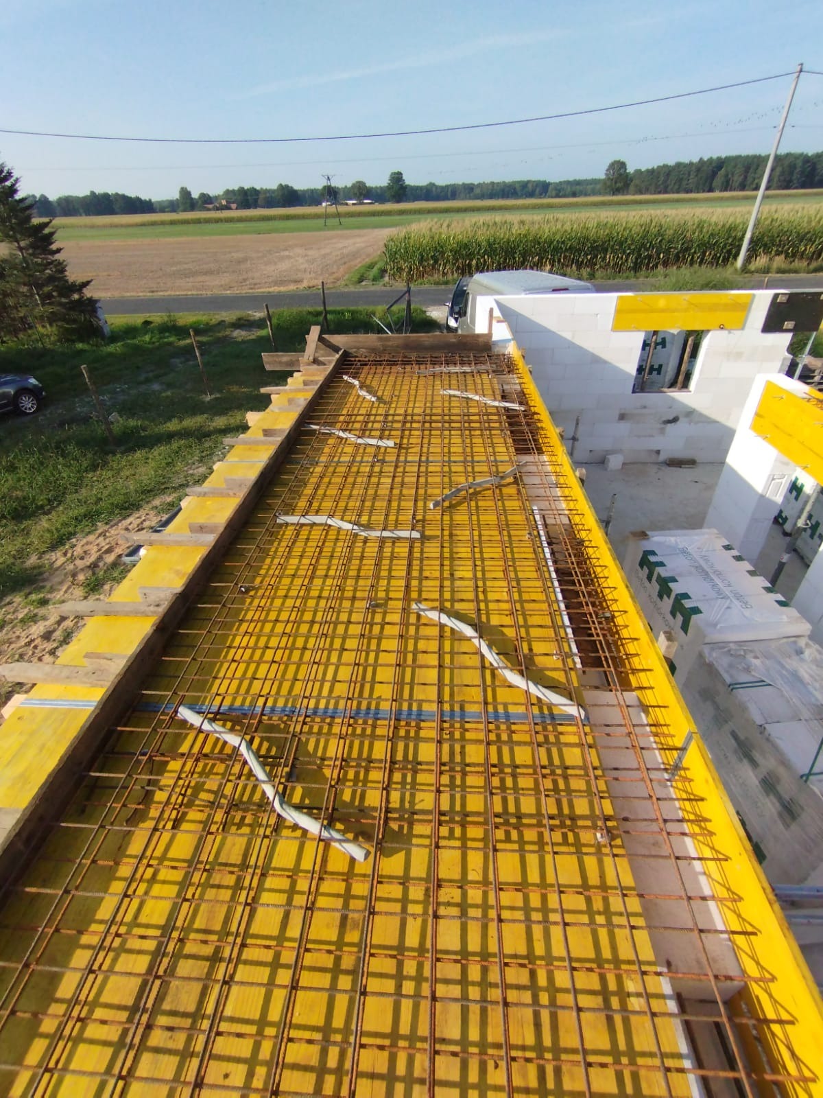 Zbrojenie stropu na żółtym szalunku, widok z góry na budowę domu jednorodzinnego z bloczków, w tle krajobraz wiejski w słoneczny dzień.