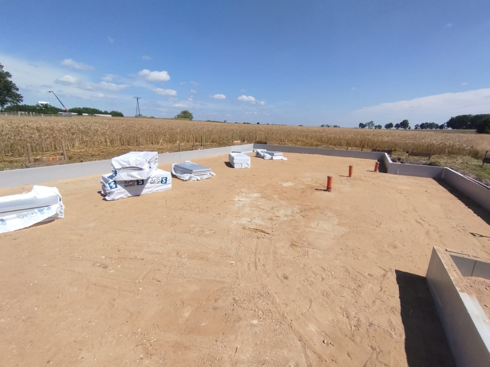 Przygotowany teren pod budowę domu z wylanymi fundamentami i ułożonymi materiałami izolacyjnymi. W tle pole zboża i błękitne niebo.