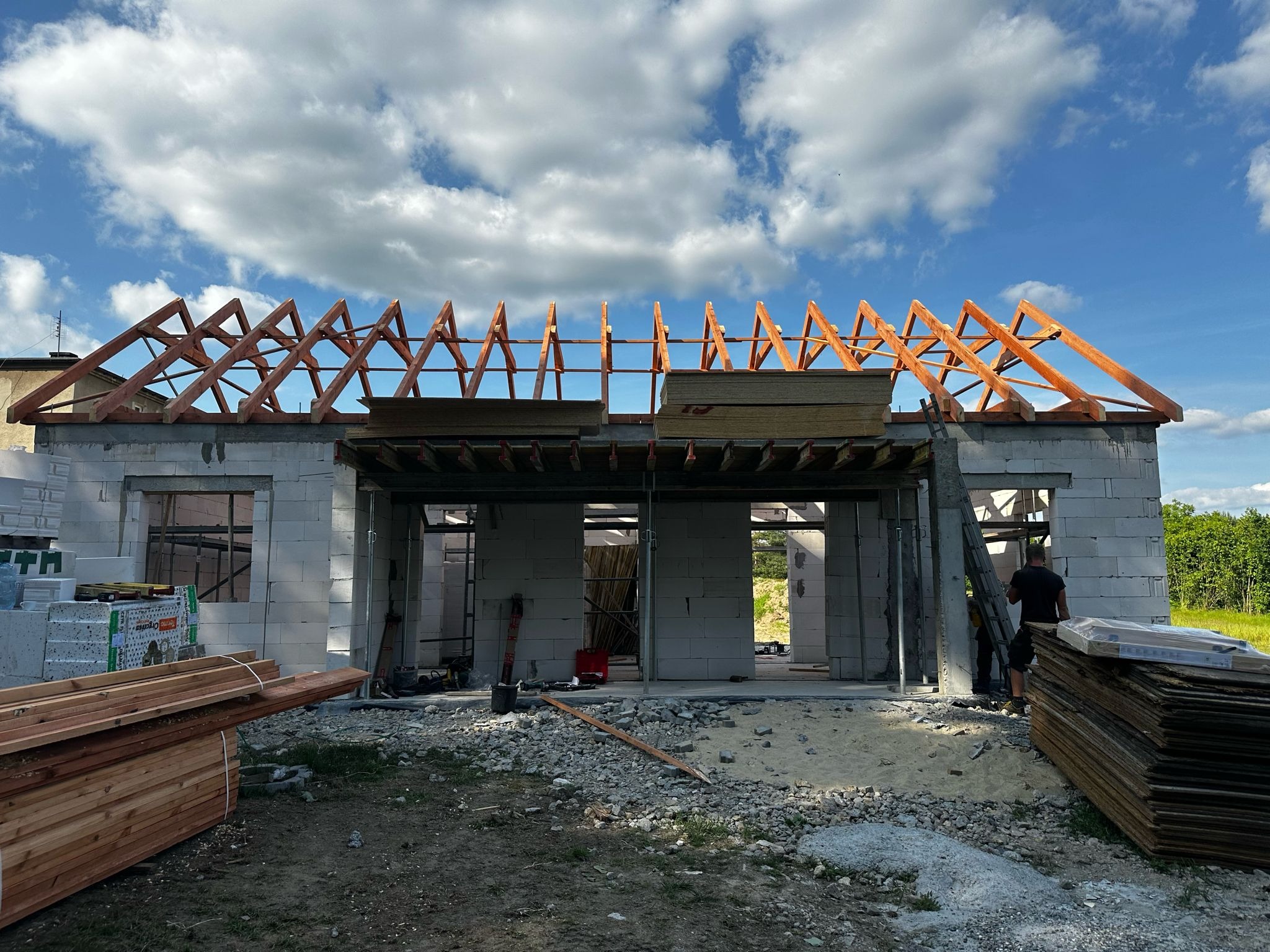 Wznoszenie domu z białych bloczków: murowane ściany, drewniana konstrukcja dachu w trakcie montażu, błękitne niebo z chmurami w tle. Widoczne materiały budowlane.