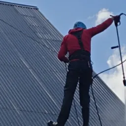 Monter w kasku, z zabezpieczeniem linowym, instaluje panele na falistym dachu z blachy, na tle błękitnego nieba z chmurami.