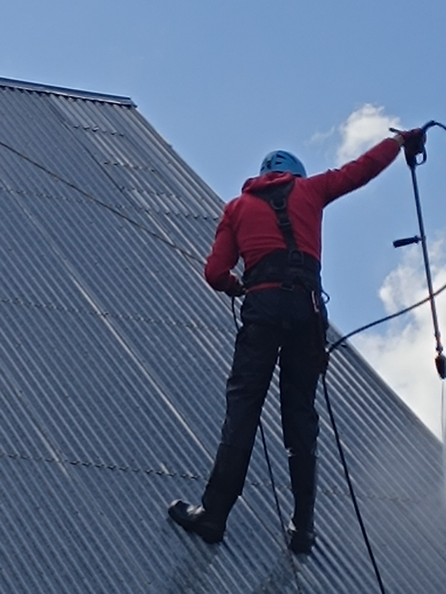 Monter w kasku, z zabezpieczeniem linowym, instaluje panele na falistym dachu z blachy, na tle błękitnego nieba z chmurami.