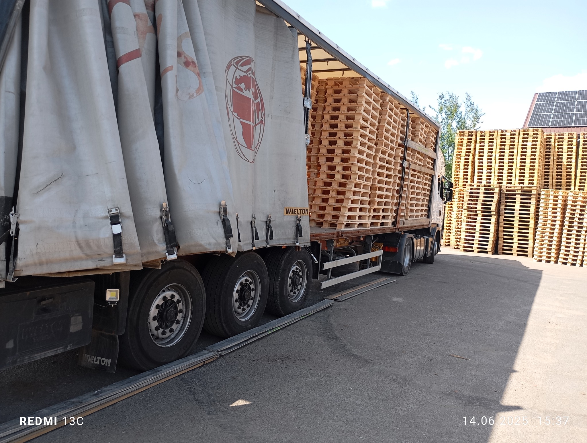 Ciężarówka marki Wielton z naczepą, załadowana paletami, zaparkowana na placu. Widoczne panele słoneczne w tle. Transport ciężarowy.