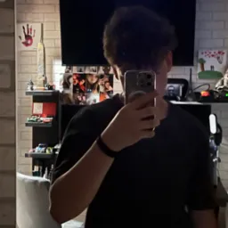 Osoba robi selfie w lustrze, w tle półki z modelami aut, zdjęcia i dekoracje na tle ściany z cegły.