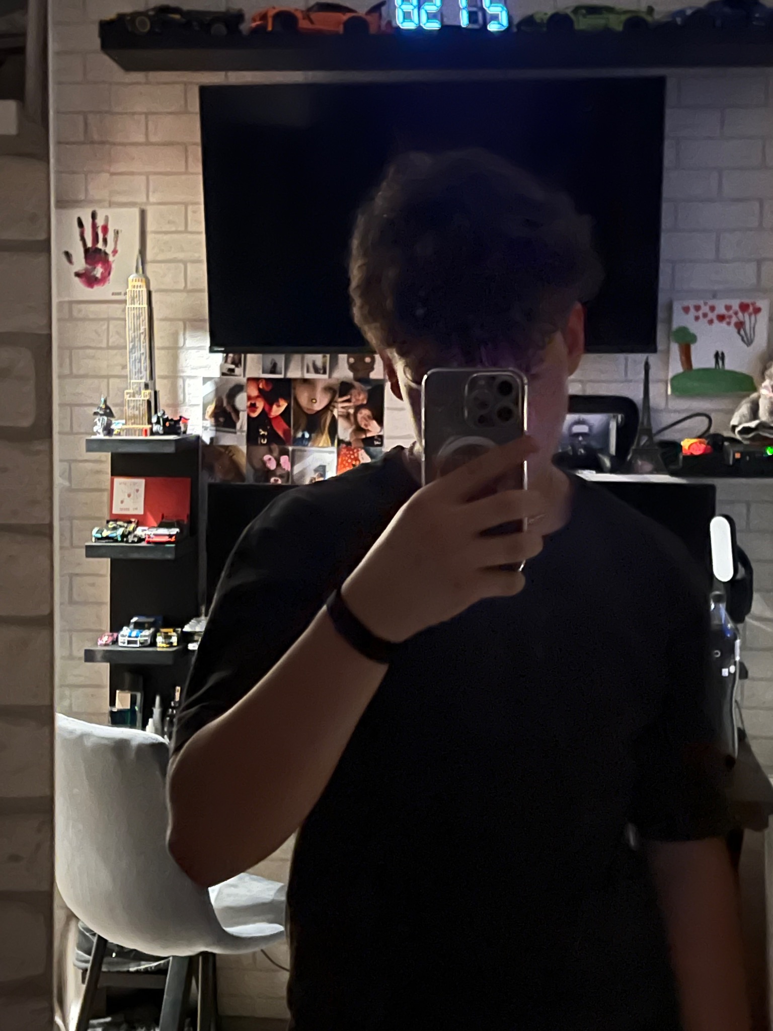 Osoba robi selfie w lustrze, w tle półki z modelami aut, zdjęcia i dekoracje na tle ściany z cegły.