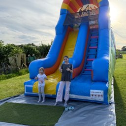 Balonowy Zamek Natalia Śremba - Dwie dziewczynki z podniesionymi kciukami na tle dmuchanej zjeżdżalni z motywem bajki. Zjeżdżalnia stoi na trawie, w tle zieleń i zachodzące słońce.