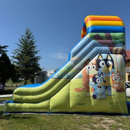 Balonowy Zamek Natalia Śremba - Kolorowa, dmuchana zjeżdżalnia z motywem bajki Bluey na trawie, w tle drzewa i budynek. Zjeżdżalnia z żółto-niebieskim ślizgiem i postaciami z bajki.