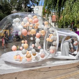Balonowy Zamek Natalia Śremba - Przezroczysta, dmuchana kula wypełniona pastelowymi balonami, ustawiona na zewnątrz. W tle budynki i zieleń, w środku widoczne osoby.