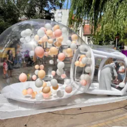 Przezroczysta, dmuchana kula wypełniona pastelowymi balonami, ustawiona na zewnątrz. W tle budynki i zieleń, w środku widoczne osoby.