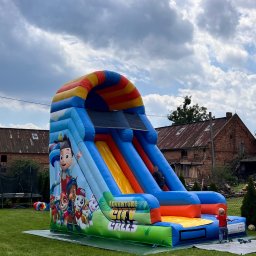 Balonowy Zamek Natalia Śremba - Kolorowa, dmuchana zjeżdżalnia z motywem 'Psi Patrol' na zielonym trawniku, z dzieckiem w czerwonej koszulce u dołu. W tle budynek gospodarczy i pochmurne niebo.