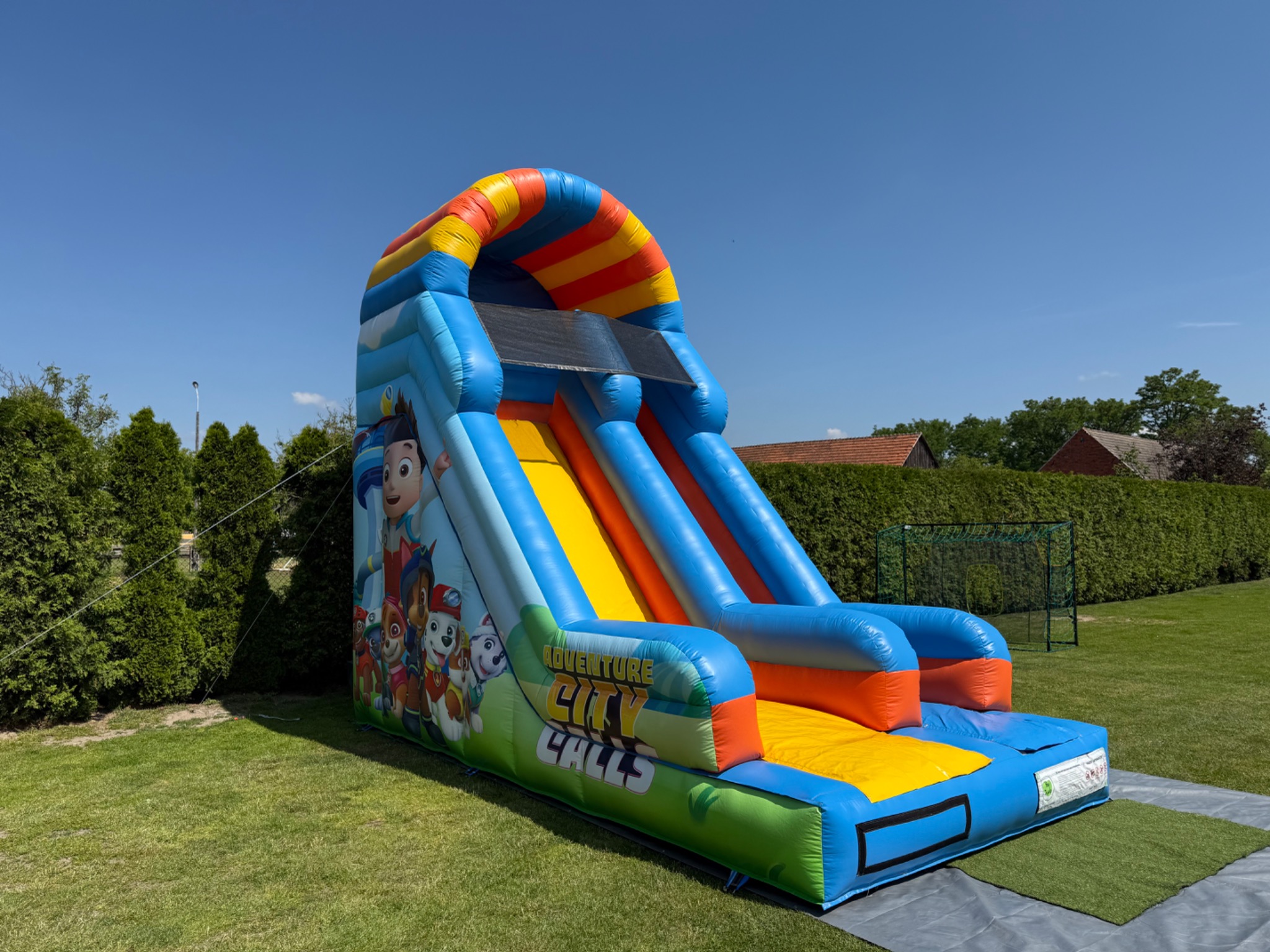 Kolorowa, dmuchana zjeżdżalnia z motywem bajki Psi Patrol, ustawiona na trawniku w ogrodzie, w słoneczny dzień. Widoczne drzewa i fragment budynku w tle.