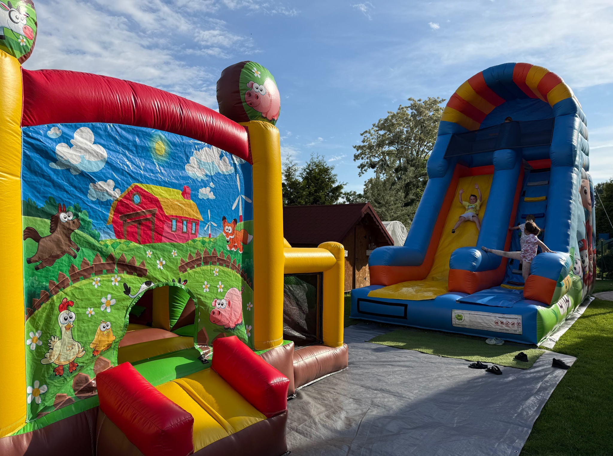Kolorowe dmuchańce z motywem farmy i zjeżdżalnią, dzieci bawią się na tle błękitnego nieba. Dmuchany zamek i zjeżdżalnia na trawie, widok z bliska.