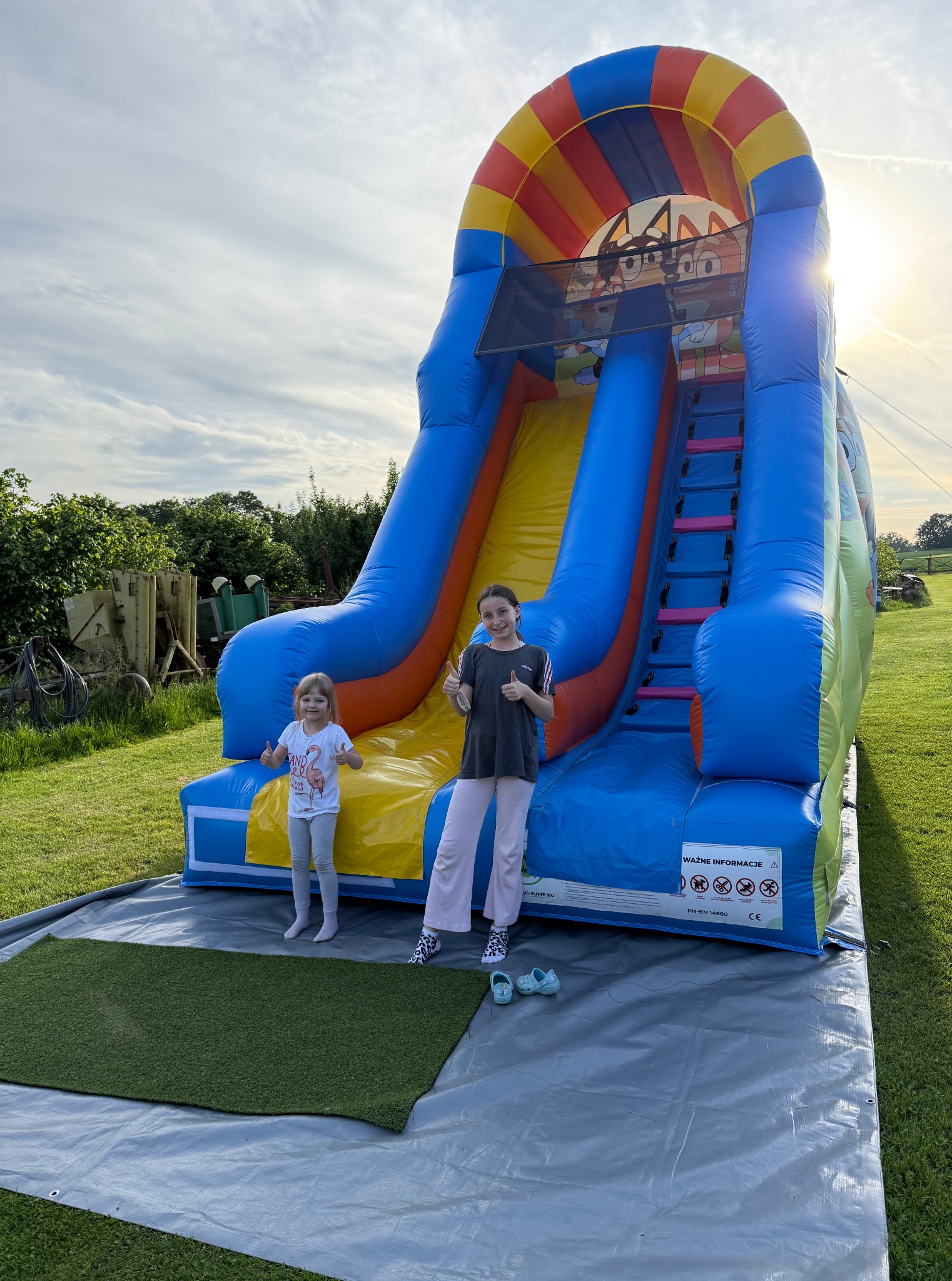 Dwie dziewczynki z podniesionymi kciukami na tle dmuchanej zjeżdżalni z motywem bajki. Zjeżdżalnia stoi na trawie, w tle zieleń i zachodzące słońce.