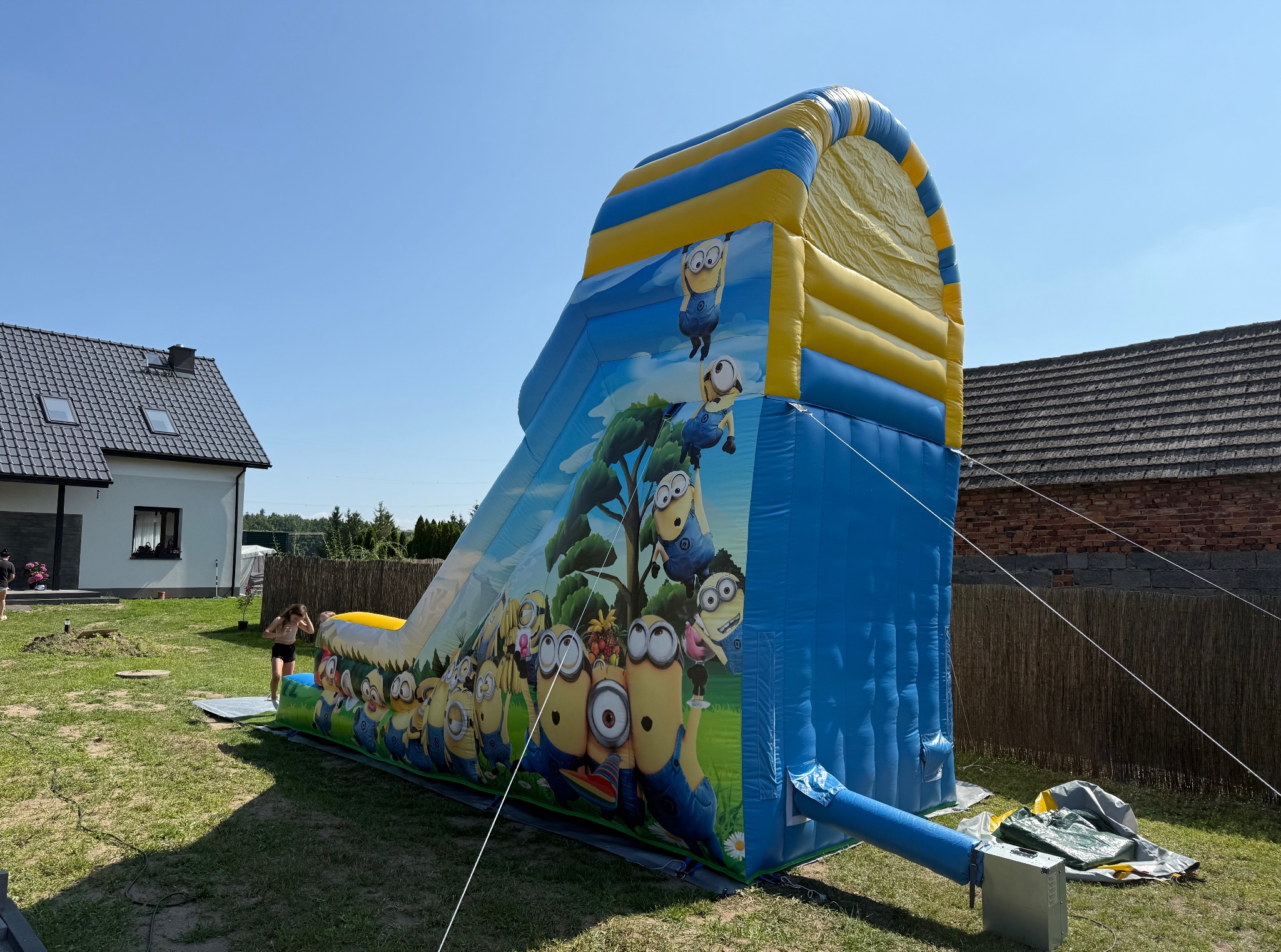 Rozłożony, kolorowy dmuchany zamek ze zjeżdżalnią z motywem Minionków na trawniku przed domem jednorodzinnym i drewnianym płotem w słoneczny dzień.