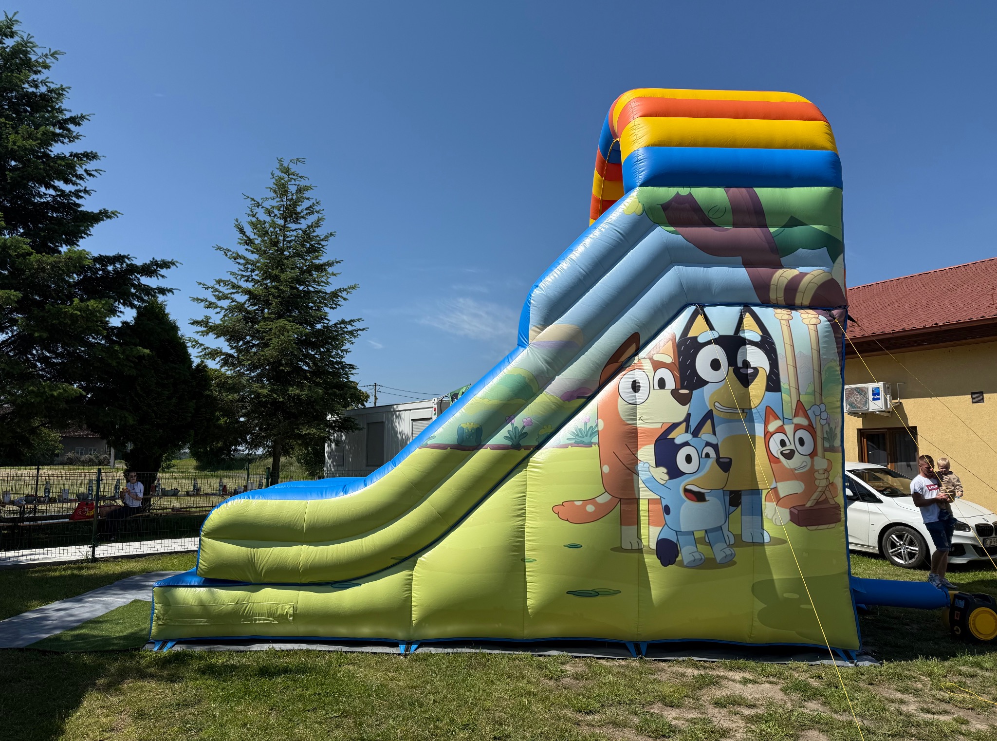 Kolorowa, dmuchana zjeżdżalnia z motywem bajki Bluey na trawie, w tle drzewa i budynek. Zjeżdżalnia z żółto-niebieskim ślizgiem i postaciami z bajki.