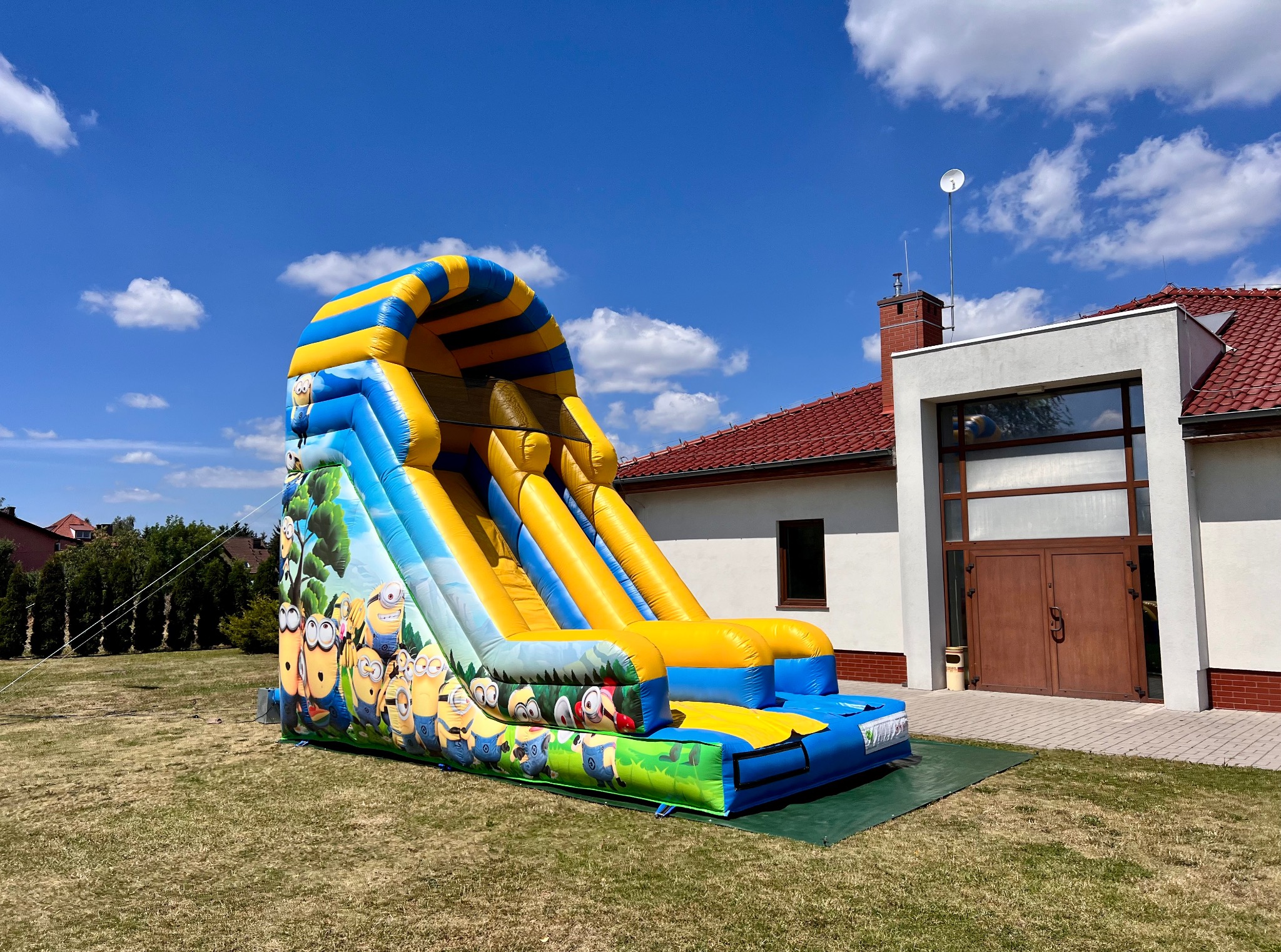 Kolorowa dmuchana zjeżdżalnia z motywem Minionków na trawie przed budynkiem w słoneczny dzień. Atrakcja na imprezy plenerowe dla dzieci.
