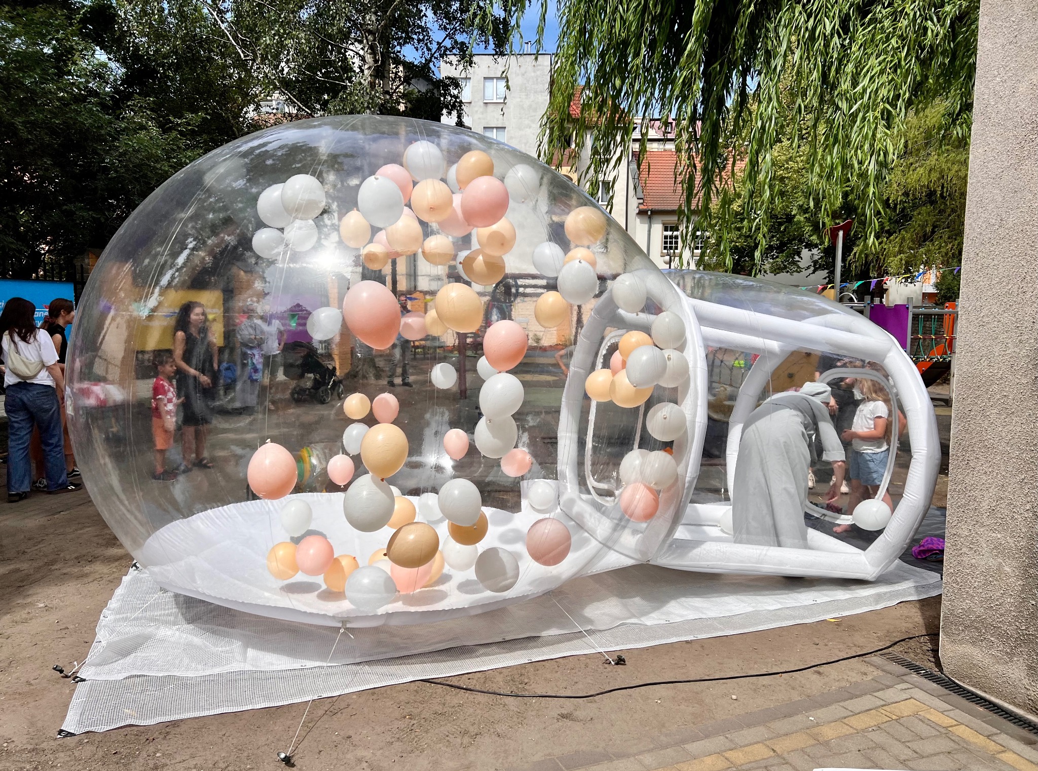 Przezroczysta, dmuchana kula wypełniona pastelowymi balonami, ustawiona na zewnątrz. W tle budynki i zieleń, w środku widoczne osoby.