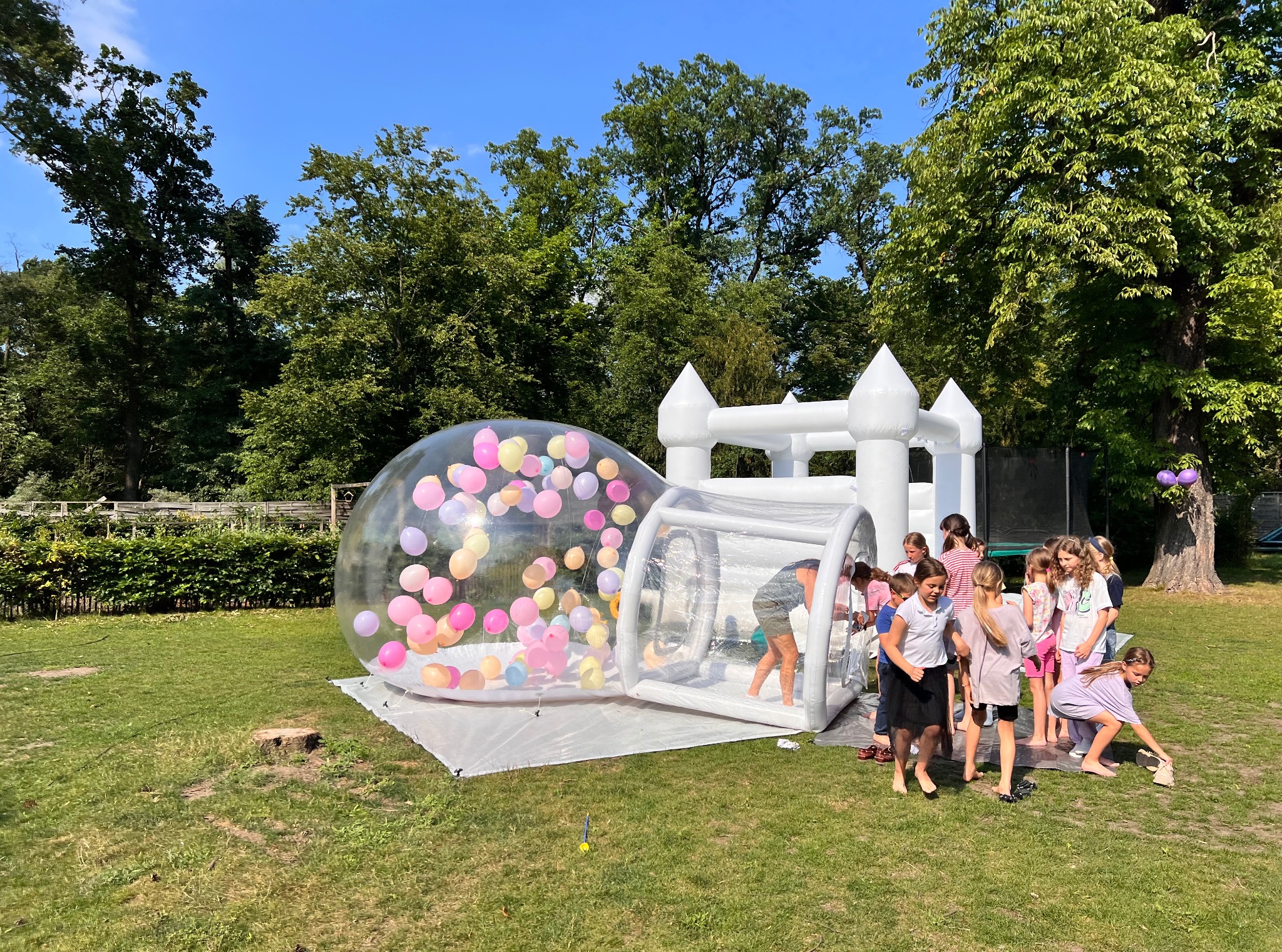 Dmuchany zamek i przezroczysta kula z balonami w Marcinowie, otoczone grupą dzieci na trawie. Atrakcja na imprezę plenerową, w tle drzewa i niebo.