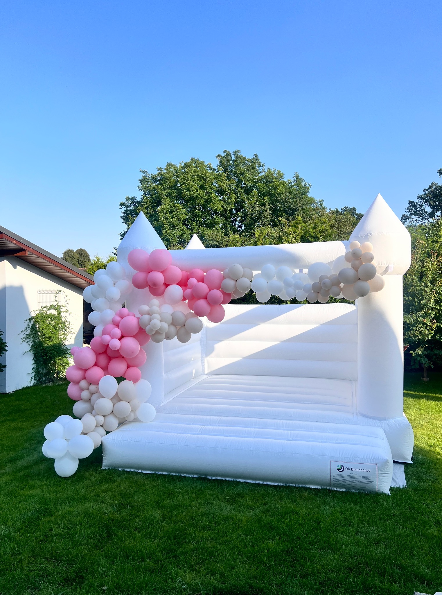 Biały dmuchany zamek, udekorowany girlandą z białych, różowych i beżowych balonów, ustawiony na trawie w ogrodzie. Błękitne niebo w tle.