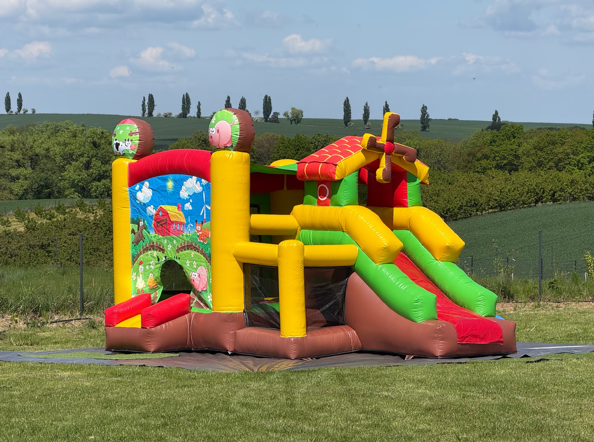 Kolorowy dmuchany zamek w kształcie farmy z dwoma zjeżdżalniami, ustawiony na trawie z widokiem na zielone wzgórza pod błękitnym niebem. Detal: dekoracje z motywami zwierząt.