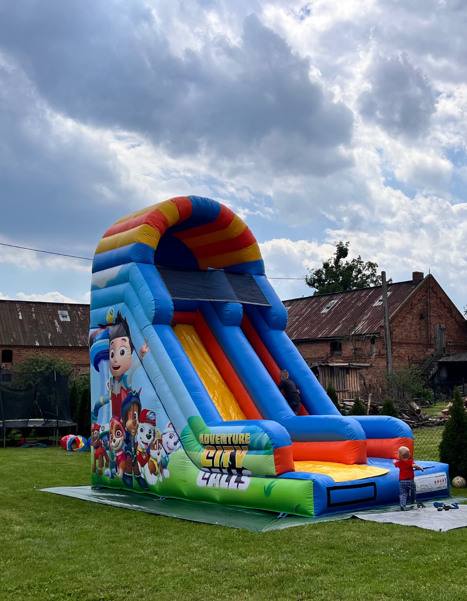 Kolorowa, dmuchana zjeżdżalnia z motywem 'Psi Patrol' na zielonym trawniku, z dzieckiem w czerwonej koszulce u dołu. W tle budynek gospodarczy i pochmurne niebo.