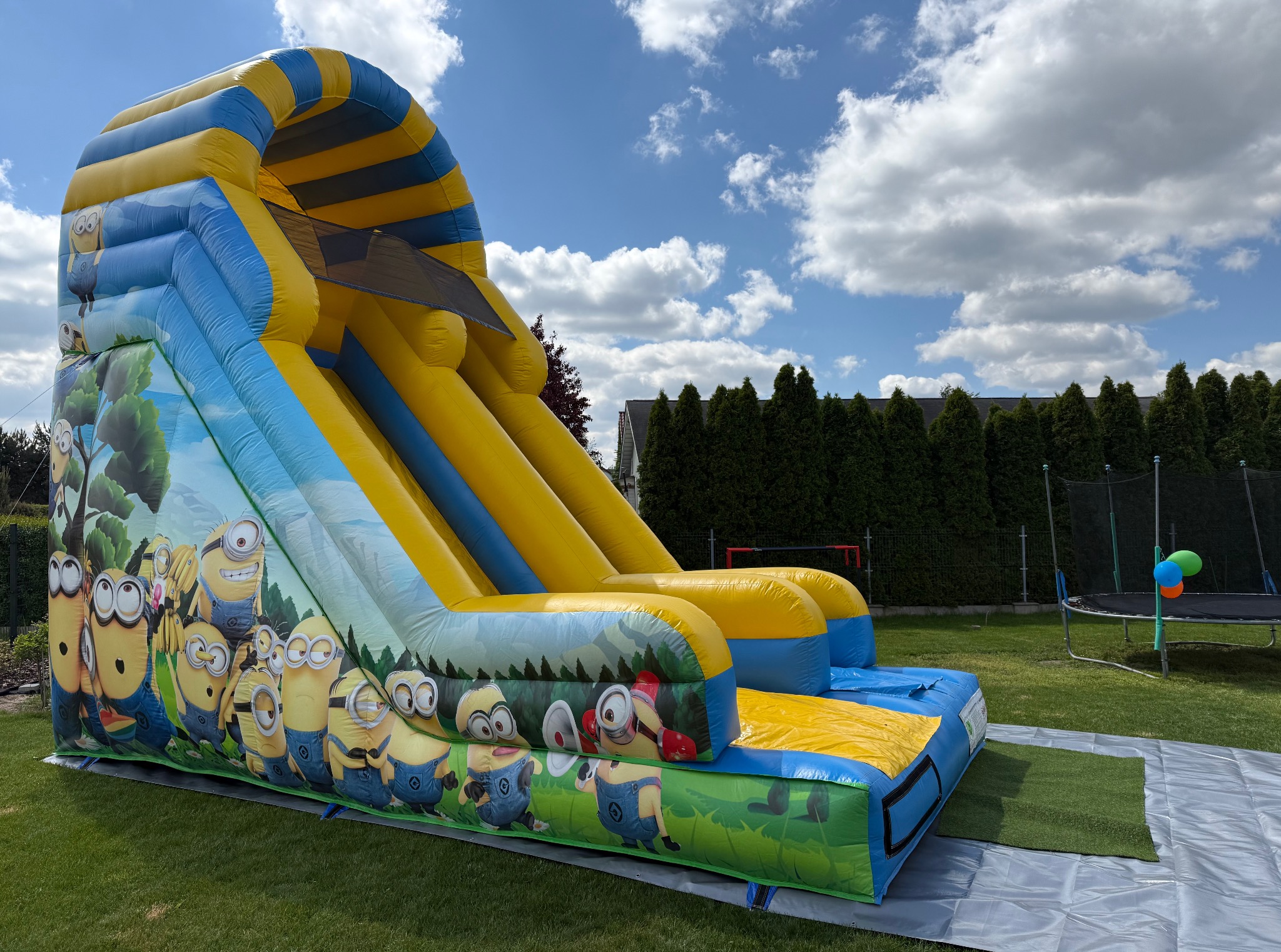 Żółto-niebieska dmuchana zjeżdżalnia z motywem Minionków na trawie, w tle zielone tuje i trampolina z balonami. Słoneczny dzień, błękitne niebo z obłokami.
